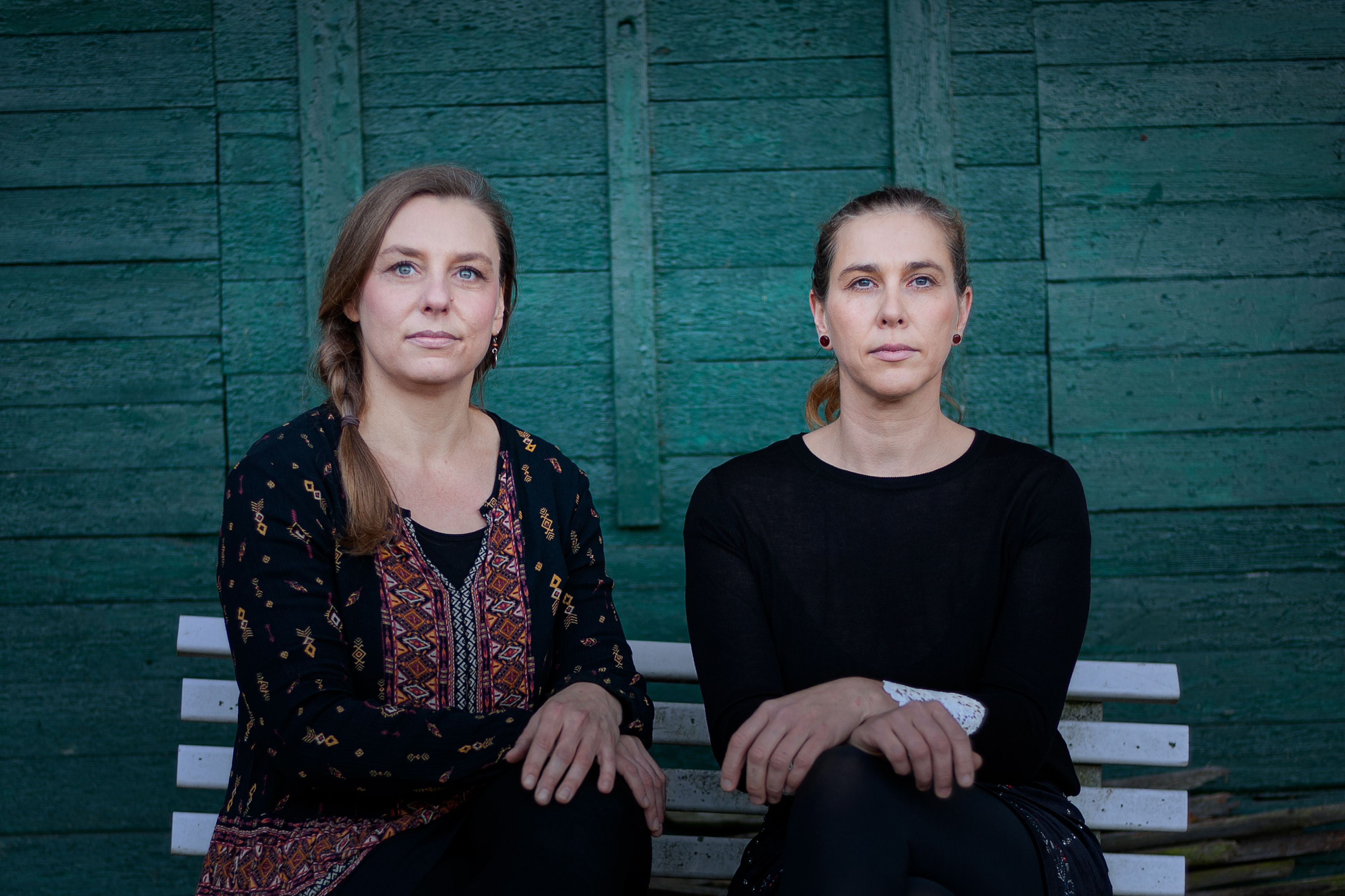 Lena und Jenny sitzen auf einer Bank vor einer dunkelgrünen Holzwand